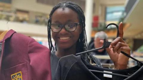 Daniella, a girl, wearing glasses, holding two blazers, a red one and a black  one on hangers in tow hands, smiling for camera.