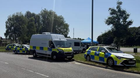 Four police vehicles are seen parked on the edge of a green space and there is a blue tent in the background