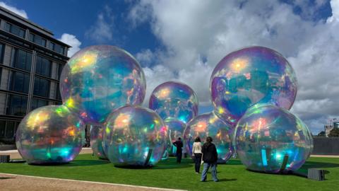 Giant bubbles that are tall. They are on green grass. There are people walking towards the bubbles with their backs turned to the camera. The sky is blue with white clouds. 