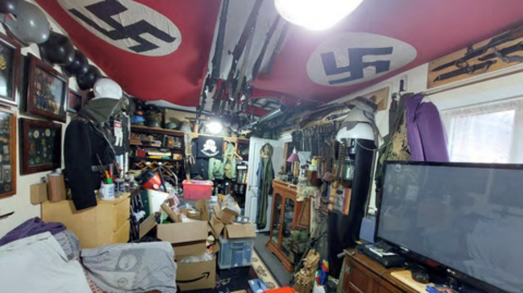 The room of a large shed with two nazi flags hung across the sealing, a TV on the right and military items on the left.