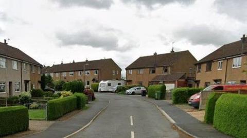 Semi-detached houses run along either side of the street, with hedges to the left and right.