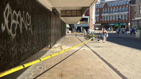 A pile of debris can be seen on the pavement underneath the canopy of a large department store. Yellow tape has cordoned the area off. People walk by. 