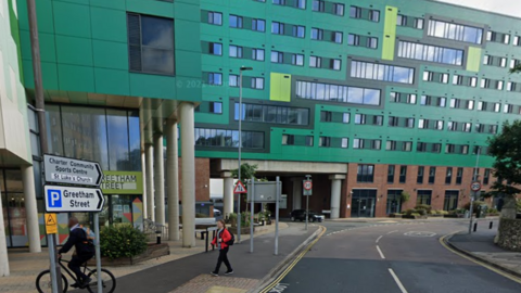 A cyclist and a pedestrian pass student halls of residence on a road with a right hand bend. The building has at least eight floors, with the top six painted green.