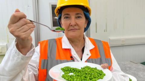Michelle is wearing a high vis jacket, a yellow hard hat, and holding a plate of peas and a spoon