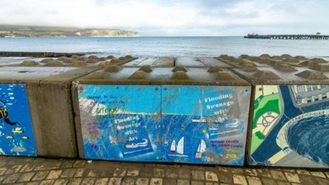 Temporary flood defences at Swanage with artwork painted on the large concrete blocks in place between the land and the sea.