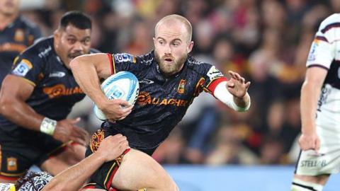 Xavier Roe is trying to escape a tackle from an opposition player. He is bald, with a short beard and is holding the rugby ball.