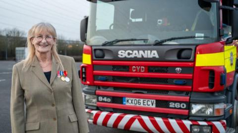 A woman in a grey suit jacket with three medals stands next to a red fire truck.