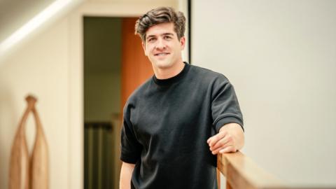 A man in a black T-shirt leans on a bannister as he looks at the camera.
