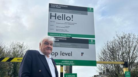 Roger Galloway is stood in front of a big green and white car park sign. He is wearing a blazer and a white shirt and is looking down directly at the camera. The car park sign has the word "Hello!" in bold text along with all the parking details as well as the Plymouth City Council logo. There are lots of trees and houses in the background. 