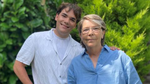 Mother and son, Jan and Rossi Griffin, standing next to each other. Jan is wearing a blue linen shirt and Rossi a white one. There are green trees in the background.