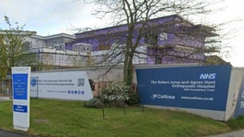 Alternative description A blue sign with the trust's name in white writing is on the right of the image. A tree and a purple coloured building are behind it on the photo.