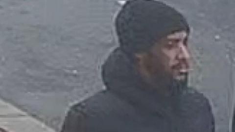 CCTV image of a man with a beard in a black coat and black woolly hat walking down the street