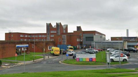 The exterior of Furness General Hospital. It is a red-brick building with cars parked at the front.