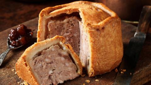 Image of pork pie sliced and served on a wooden board, with a teaspoon on cranberry sauce next to it.