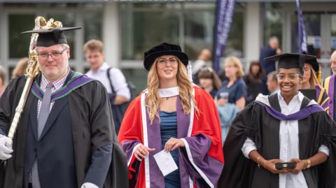 Jessica-Jane Applegate smiles at the camera as she walks with two university staff. She is wearing a red and purple robe and black hat. She has long blonde hair. She is in between a woman in black robes and a man wearing black robes and carrying a gold ceremonial mace. 