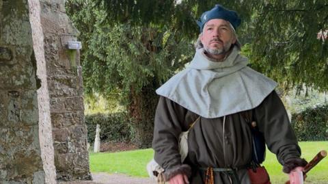 A man dressed in medieval attire walking past a church building with grass and graves seen behind. A large tree is also covering the skyline.