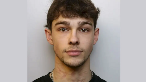 Mathew McGrath looking straight into the camera in his mugshot photo. He has curly brown hair, thick eyebrows and a thin beard and moustache. He wears a black t-shirt and a thick silver chain.