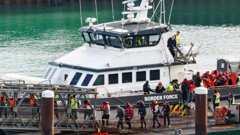 A border force vessel carries asylum seekers into port at Dover. A line of men waering life vests can be seen disembarking the vessel and walking in a line. the boat says "Border Force" along the side