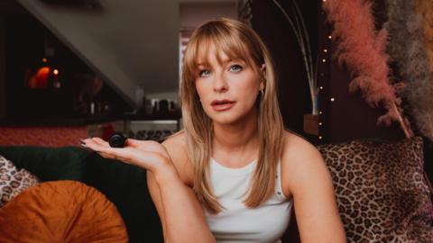 A blonde woman with a fringe looking directly at the camera whilst holding a small black spy camera in her right hand. She's also wearing a white top.
