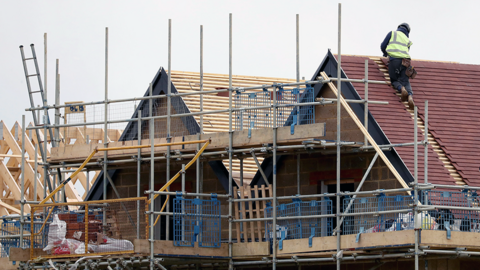 A generic image of a building site, with a builder in a high-vis jacket perched on the roof of a house. Scaffolding surrounds two properties, with a third in the background on the left