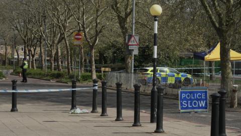 Blue police tape is attached around black bollards, with a police car seen in the background.