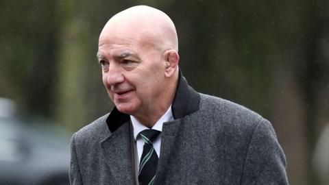 A man with a bald head, wearing a grey coat with a black collar. He is wearing a white shirt and a black tie with green and white stripes. He is looking towards the left hand side of the picture.