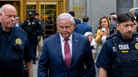 Former US Senator Bob Menendez pictured outside of a federal court in New York. He is flanked by police officers and is wearing a dark blue pin-stripped suit and red tie. Reporters are in the background. 