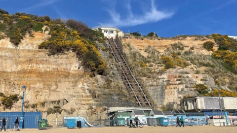 Sandy edge of beach with metal fencing, line of bins and shipping container with bare cliff seen after fall.