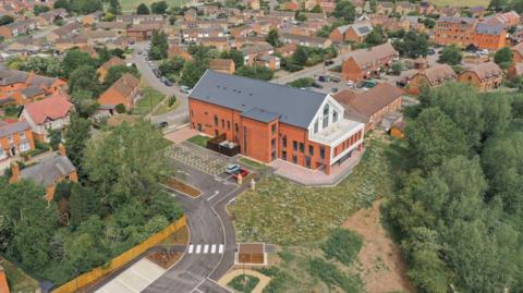 An aerial view of a modern two-storey red brick building with a dark grey roof and a distinctive white-framed glass gable at one end. The building sits in a residential area surrounded by houses, with a small car park, landscaped entrance and green countryside stretching into the distance beyond the town. The hospital appears newly built, with neat road markings and fresh planting around the site.