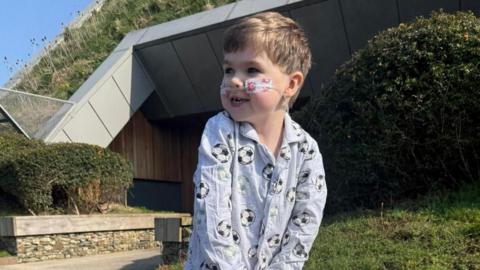 Oliver stand outside wearing grey pyjamas with footballs printed on them. He is stood on a path and surrounded by grass
