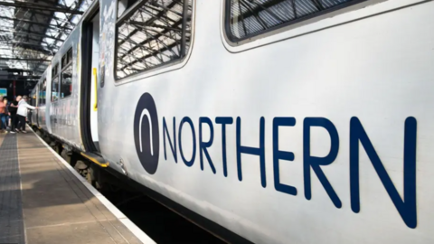 A wide shot of the side of a Northern train. It is stationary at the platform.