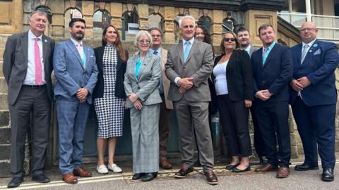 11 people in suits stood infront of grey stone building