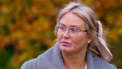 Julie Ann McIlwaine - a woman with blonde hair which is tied back with a light pink bow looks to her right as she is walking. She is wearing silver frame glasses and a grey collared coat. In the background are blurred orange and green trees.