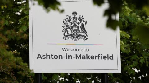 A white sign with black writing reading 'Welcome to Ashton-in-Makerfield'