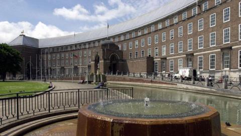 Bristol City Hall at College Green. It is a long and curved brick building with four floors, facing a large grassy area. There is a small fountain outside and flags flying on poles.