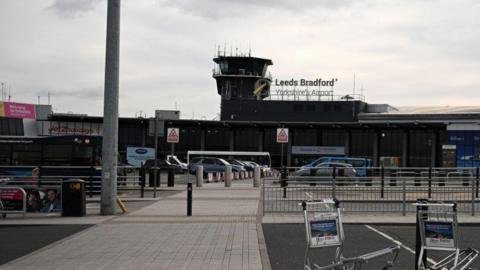 An external view of Leeds Bradford Airport. Luggage trolleys and parked cars can be seen in the airport's car park.