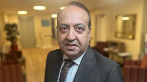 Zuffar Haq wearing a dark grey suit, white and blue striped shirt and dark tie.