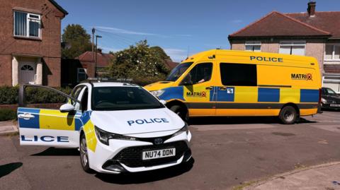 A police car and police van parked in the road on Ayton Crescent. The car has a white livery while the van's is yellow. 