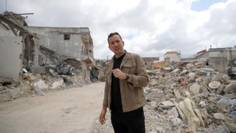 Hugo Bachega speaking in front of buildings reduced to rubble in southern Lebanon