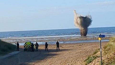 A wide picture of Ainsdale Beach as the WW2 bomb is exploded underwater. Smoke from the detonation can be see above the water, and officers are standing to watch.