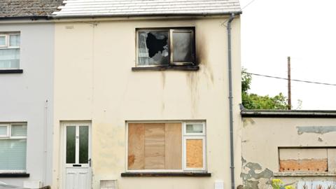 A cream coloured house with boarded windows and an upstairs window that is smashed and burned. There are burn marks down the wall.