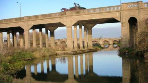 A stone viaduct above a river. Its reflection can be seen in the water. 