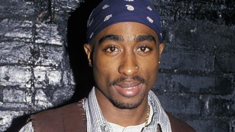 Tupac Shakur standing against a black brick wall in 1994