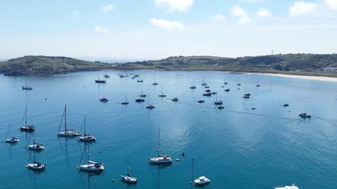 Image shows yachts on calm blue waters with golden sand and green coastline of the island of Alderney.