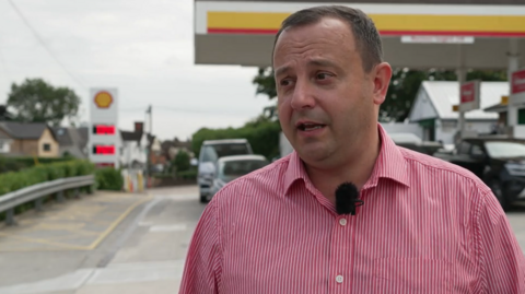Shell garage owner Goran Raven, a man with dark hair, wearing a red and white striped shirt, standing in front of the forecourt