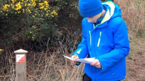 A boy in a blue raincoat and wool hat holding a paper map, which he is looking down to read. He is standing next to a wooden post with a white and orange orienteering marker.