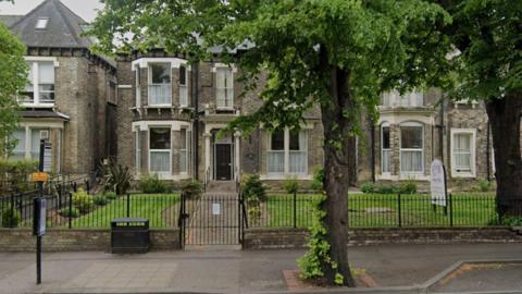 A two-storey grey brick building with a dark-coloured front door and bay windows on either side. There is a small black-fenced garden with green grass in front of the building. There is a path with a large tree in front of the garden. 
