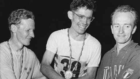 Three men wearing medals and athletics gear holding hands together in a black and white photograph. All the men are smiling, particularly the man in the centre of the image.