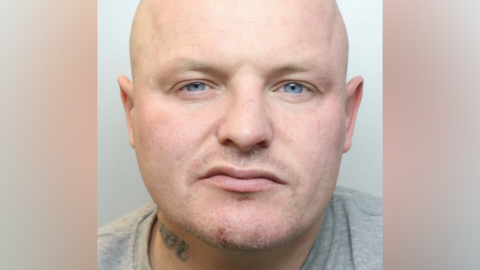 A mugshot of a bald man, wearing a grey top, scowling at the camera. A tattoo is partially visible on the right of his neck.