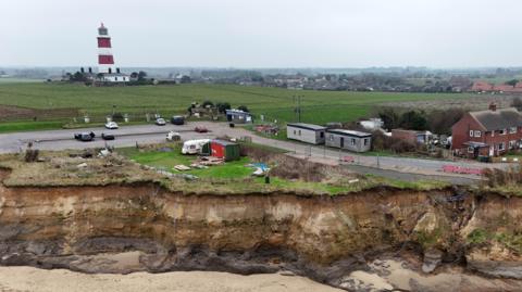 An aerial shot looking from above the sea towards the cliffs at Happisburgh. There is fencing on the road leading to the car park, preventing access to the crumbling cliffs. The lighthouse is in the background.